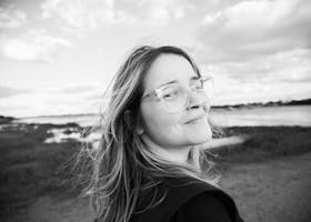 A smiling woman with glasses enjoys a breezy day at the beach in black and white.